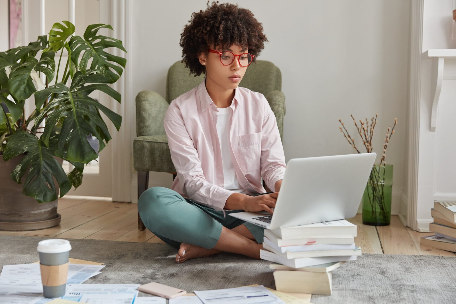Black businesswoman keyboards on laptop computer, makes diploma paper, uses laptop which stands on pile of books, drinks takeaway coffee, sits crossed legs on floor, busy working. Studying concept